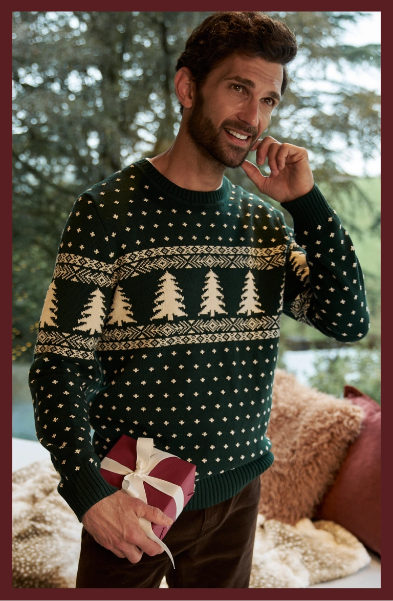 Mann trägt einen dunkelgrünen Weihnachtspullover mit Tannenbaummuster und hält ein Geschenk
